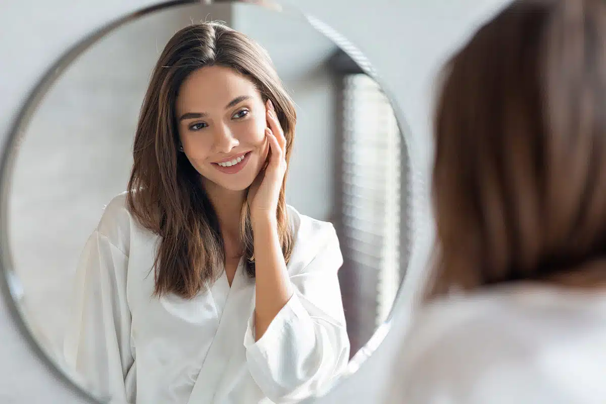 A young female patient looking at herself in the mirror admiring the results of her Facial Fat Transfer.