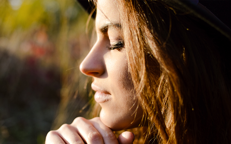 A profile shot of a woman in soft sunlight, resting her chin on her hand, with a focus on the smooth contours of her nose and facial features.