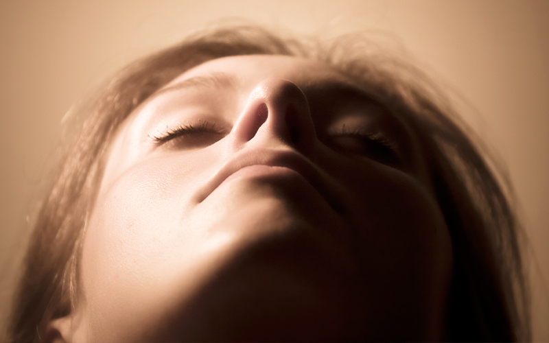 A low-angle close-up image of a woman with her eyes closed, emphasizing the shape of her nose and soft facial features under warm lighting.