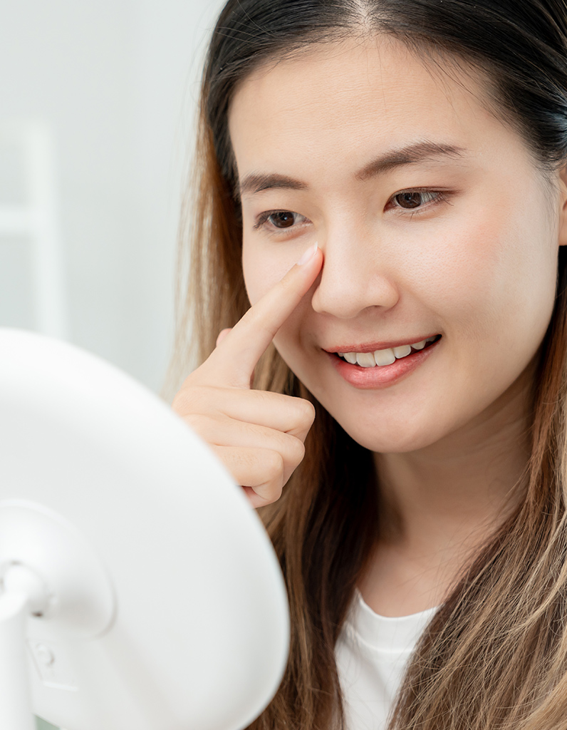 Woman touching nasal bridge while looking in mirror, representing rhinoplasty recovery and gradual reduction of swelling.