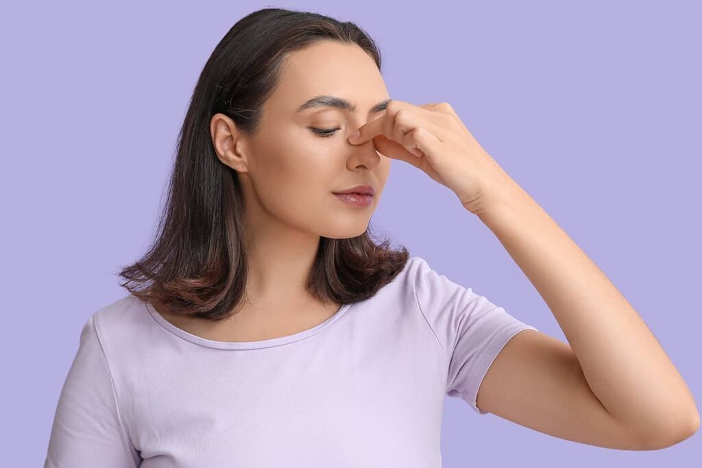 A close-up side view of a woman with her eyes closed, gently pinching the bridge of her nose against a light purple background.