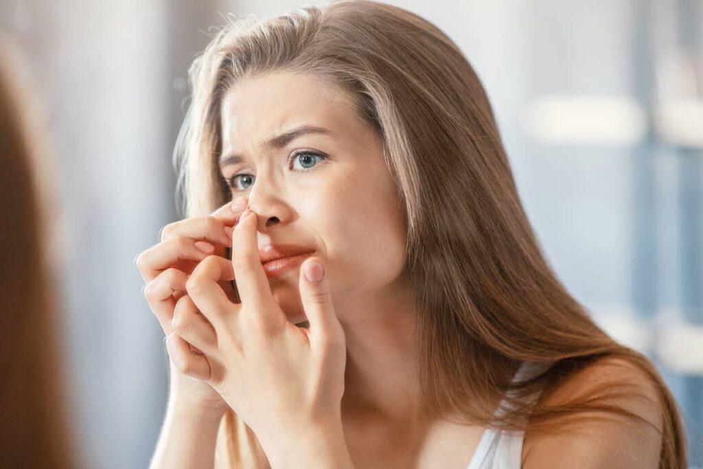 A woman looking into a mirror with a focused expression as she uses both hands to examine the symmetry of her nose.