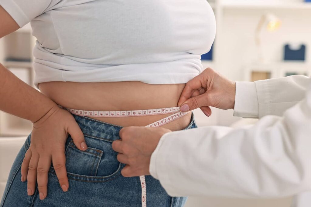 A side view of a doctor in a white lab coat using a tape measure to take baseline measurements of a patient's abdomen.
