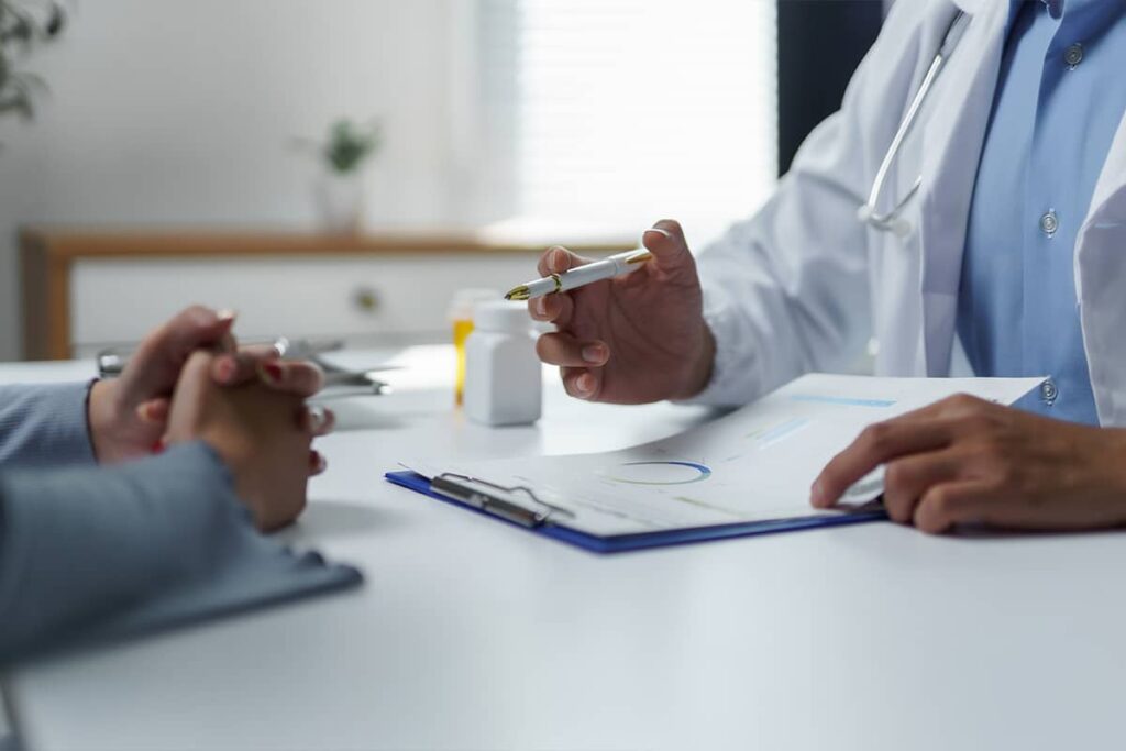 A close-up view showing a doctor’s hand with a pen pointing to specific diagrams and text on a medical form while a patient sits with their hands clasped.
