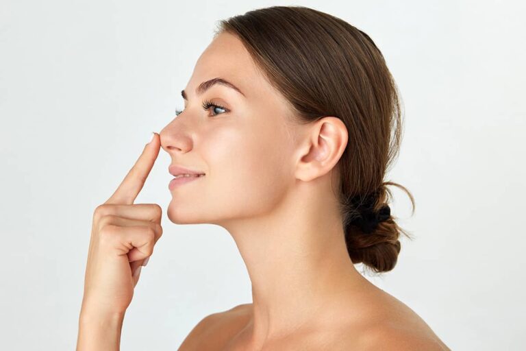 A side profile view of a brunette woman with her hair in a low bun, smiling while gently pointing her index finger toward the tip of her nose.