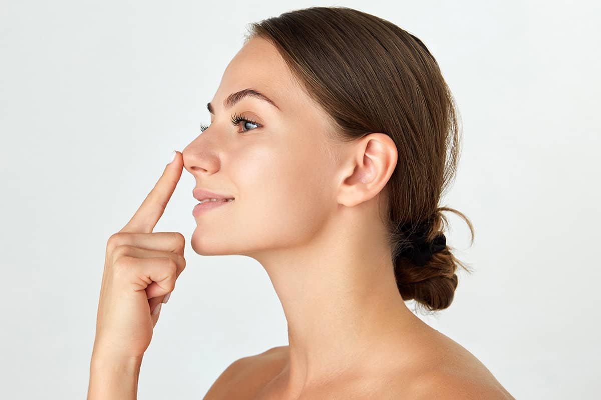 A side profile view of a brunette woman with her hair in a low bun, smiling while gently pointing her index finger toward the tip of her nose.