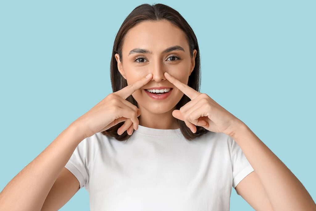 A front-facing portrait of a smiling woman in a white t-shirt using both index fingers to point directly at her nostrils against a blue background.