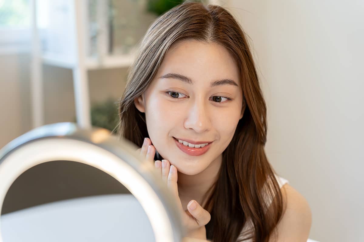 A young woman with brown hair looking at her reflection in a circular, lighted vanity mirror while gently checking the skin on her jawline.